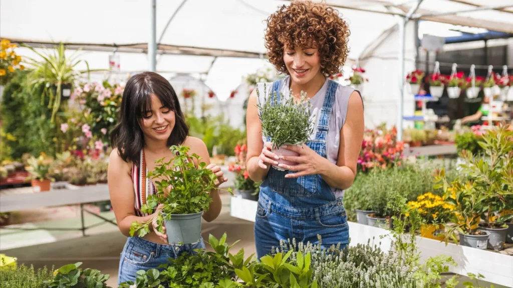 ocal garden center outdoor living inspiration - Redmonds Garden Center Two women in a plant shop holding potted herbs and smiling at each other, surrounded by greenery and colorful flowers.